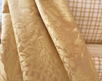 May include: Close-up of a folded, golden-yellow fabric with a detailed floral and basketweave pattern. The fabric has a luxurious sheen and is draped over a plaid patterned surface, suggesting upholstery or drapery.