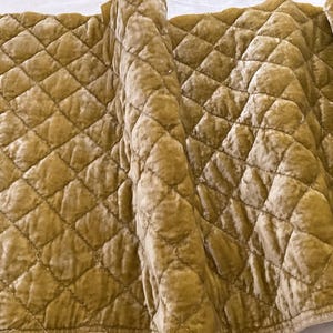 May include: Close-up of a quilted fabric in a warm, golden-yellow hue. The fabric features a diamond pattern, with each diamond slightly raised, creating a textured surface. The material appears soft and plush, with a subtle sheen.