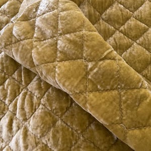 May include: Close-up of a section of quilted fabric in a golden-yellow color. The fabric has a soft, velvety texture and a diamond pattern created by the stitching. The fabric is folded over, showing the texture.