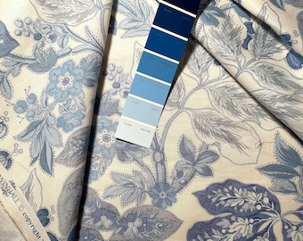 May include: Cream-colored fabric with a blue floral pattern. The design features large flowers, leaves, and vines in shades of blue and gray. A blue color swatch is included for color comparison. The fabric is folded, showing the pattern's repeat.