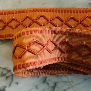 May include: Close-up of two pieces of orange ribbon with a diamond pattern. The ribbon has a textured appearance with a repeating design of stitched diamonds in a darker orange color. The edges are also detailed with a stitched border.