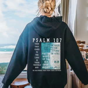 May include: Black hoodie with a white graphic print of Psalm 107:28-29. The graphic features a cross and waves with the text "He made the storm be still and the waves of the sea were hushed".
