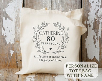 May include: A white canvas tote bag with a black design featuring a wreath with the text "Catherine 80 Years Young" and the words "A lifetime of memories, a legacy of love." The text "PERSONALIZE TOTE BAG WITH NAME" is also visible.