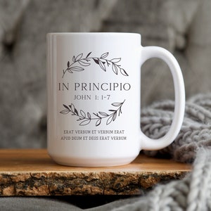 May include: White ceramic mug with a black design featuring a floral wreath and the text "IN PRINCIPIO JOHN 1: 1-7 ERAT VERBUM ET VERBUM ERAT APUD DEUM ET DEUS ERAT VERBUM".