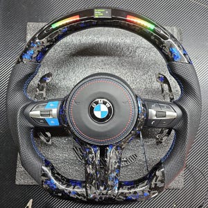 K&ouml;nnte beinhalten: Ein schwarzes Lederlenkrad mit blauen und silbernen Akzenten. Das Lenkrad hat ein Carbonfaser-Muster und ein BMW-Logo in der Mitte. Das Lenkrad verf&uuml;gt au&szlig;erdem &uuml;ber Bedienelemente f&uuml;r das Audiosystem des Fahrzeugs und die Geschwindigkeitsregelung.