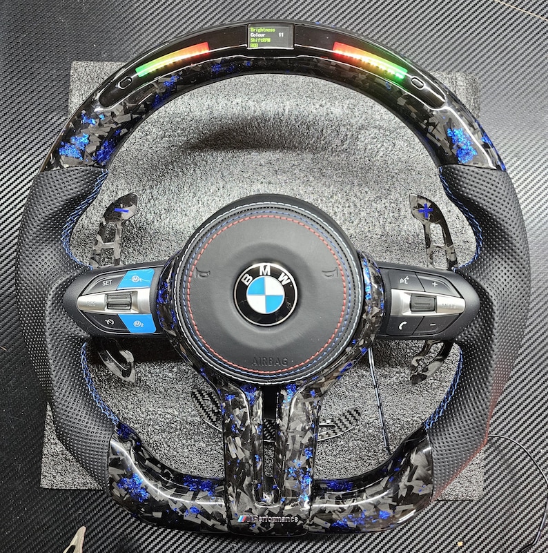 K&ouml;nnte beinhalten: Ein schwarzes Lederlenkrad mit blauen und silbernen Akzenten. Das Lenkrad hat ein Carbonfaser-Muster und ein BMW-Logo in der Mitte. Das Lenkrad verf&uuml;gt au&szlig;erdem &uuml;ber Bedienelemente f&uuml;r das Audiosystem des Fahrzeugs und die Geschwindigkeitsregelung.