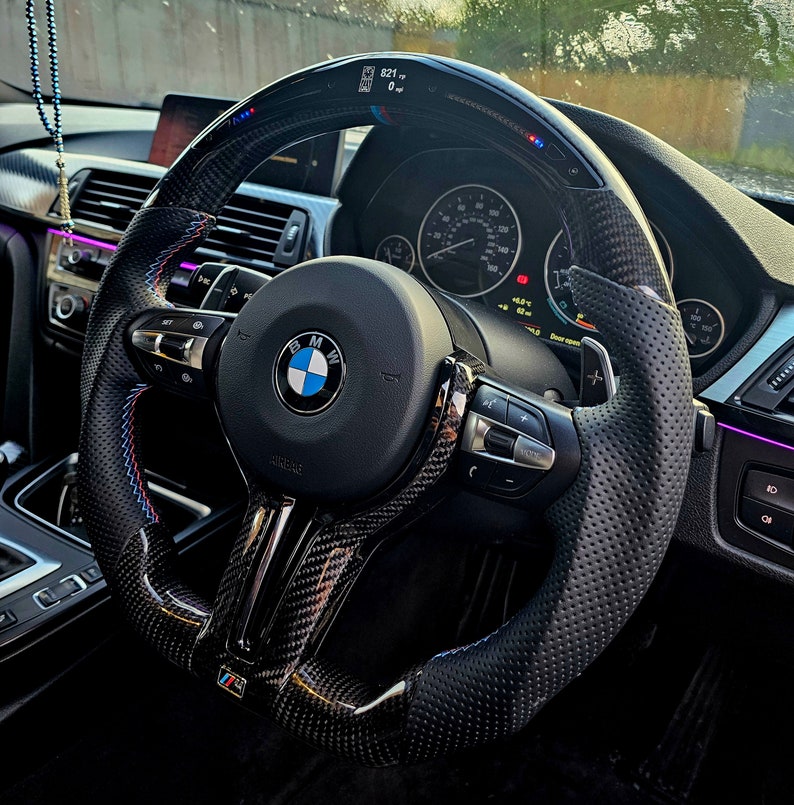 May include: A black leather steering wheel with a carbon fiber trim and a BMW logo. The steering wheel has a digital display showing the speed and other information. The steering wheel has paddle shifters on the back.