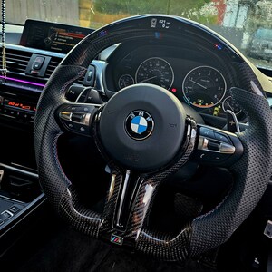 May include: A black leather steering wheel with carbon fiber accents and blue and red stitching. The steering wheel has a BMW logo and the words "Airbag" and "M" printed on it.
