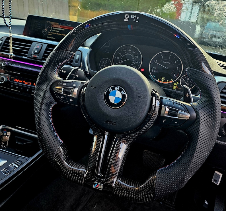 May include: A black leather steering wheel with carbon fiber accents and blue and red stitching. The steering wheel has a BMW logo and the words "Airbag" and "M" printed on it.
