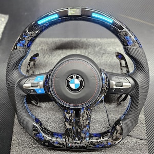 K&ouml;nnte beinhalten: Ein schwarzes Lederlenkrad mit blauen N&auml;hten und einem Karbonfaser-Muster. Das Lenkrad hat einen blauen LED-Lichtstreifen um den Rand und ein BMW-Logo in der Mitte. Das Lenkrad verf&uuml;gt au&szlig;erdem &uuml;ber Bedienelemente f&uuml;r das Audiosystem des Fahrzeugs und die Geschwindigkeitsregelung.