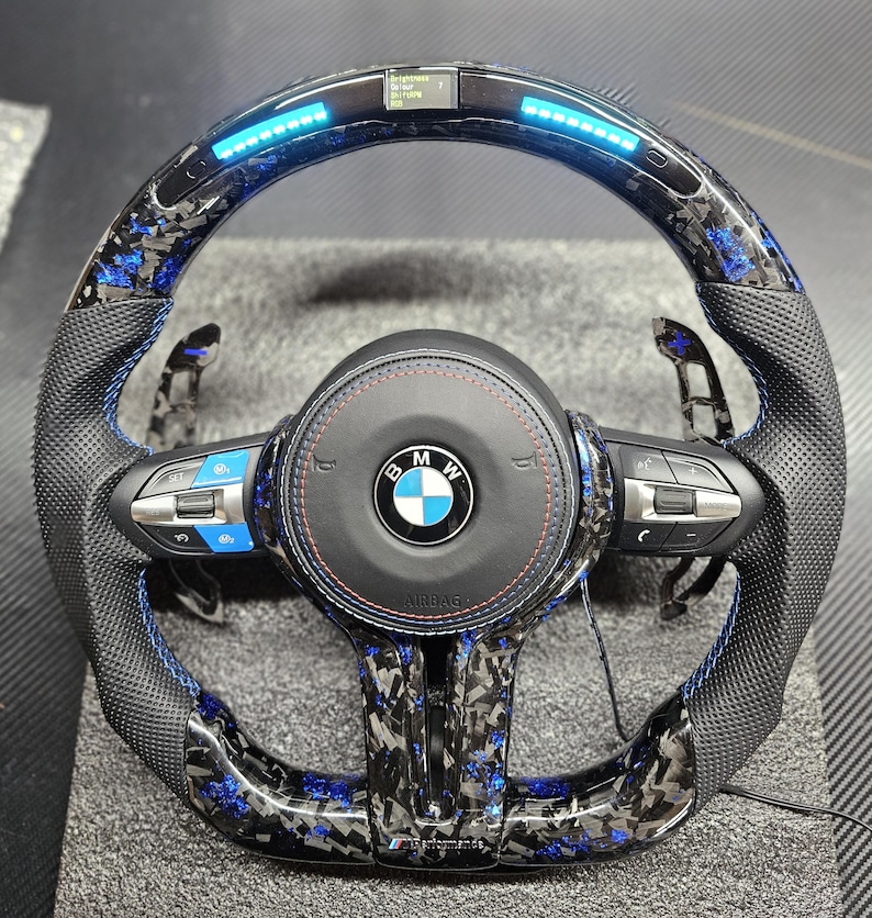 K&ouml;nnte beinhalten: Ein schwarzes Lederlenkrad mit blauen N&auml;hten und einem Karbonfaser-Muster. Das Lenkrad hat einen blauen LED-Lichtstreifen um den Rand und ein BMW-Logo in der Mitte. Das Lenkrad verf&uuml;gt au&szlig;erdem &uuml;ber Bedienelemente f&uuml;r das Audiosystem des Fahrzeugs und die Geschwindigkeitsregelung.
