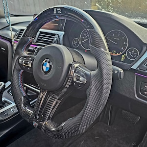 May include: A black leather steering wheel with a carbon fiber trim and a perforated surface. The steering wheel has a BMW logo and a variety of buttons and controls.