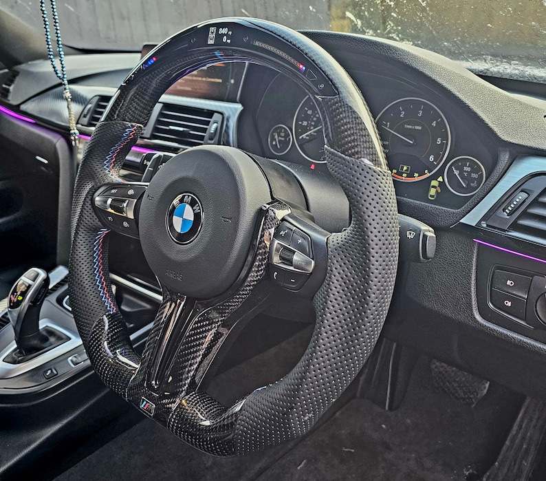 May include: A black leather steering wheel with a carbon fiber trim and a perforated surface. The steering wheel has a BMW logo and a variety of buttons and controls.