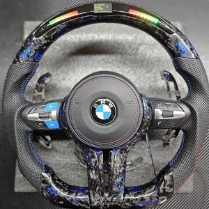 K&ouml;nnte beinhalten: Ein schwarzes Lederlenkrad mit blauen N&auml;hten und einem Karbonfaser-Muster. Das Lenkrad tr&auml;gt das BMW-Logo und den Schriftzug "Airbag". Das Lenkrad verf&uuml;gt au&szlig;erdem &uuml;ber eine Vielzahl von Kn&ouml;pfen und Bedienelementen.