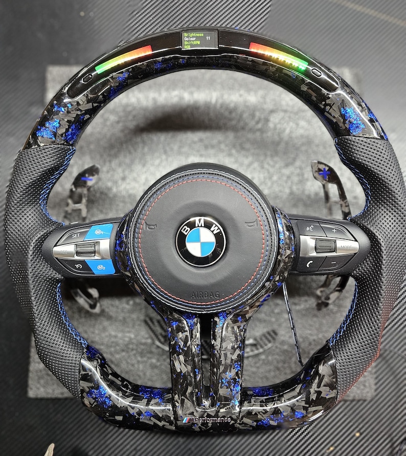 K&ouml;nnte beinhalten: Ein schwarzes Lederlenkrad mit blauen N&auml;hten und einem Karbonfaser-Muster. Das Lenkrad tr&auml;gt das BMW-Logo und den Schriftzug "Airbag". Das Lenkrad verf&uuml;gt au&szlig;erdem &uuml;ber eine Vielzahl von Kn&ouml;pfen und Bedienelementen.