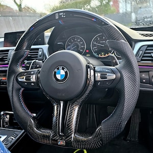 May include: A black leather steering wheel with carbon fiber accents and a perforated grip. The steering wheel has a BMW logo and the text "Airbag" printed on it. The steering wheel also has controls for the car's audio system and cruise control.