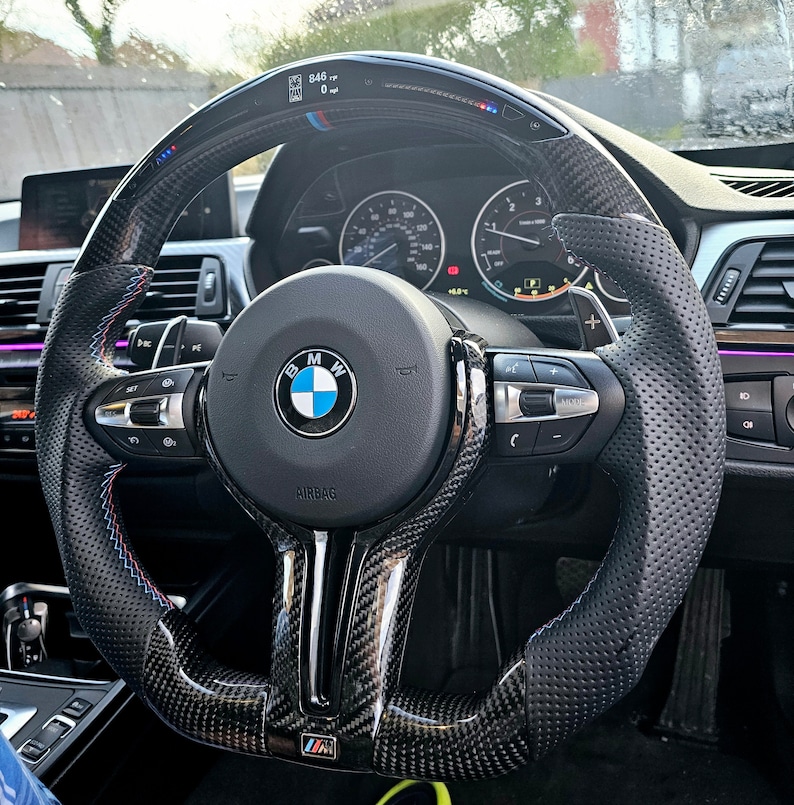 May include: A black leather steering wheel with carbon fiber accents and a perforated grip. The steering wheel has a BMW logo and the text "Airbag" printed on it. The steering wheel also has controls for the car's audio system and cruise control.
