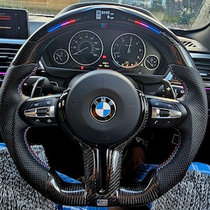 May include: A black leather steering wheel with a carbon fiber trim and blue and red stitching. The steering wheel has a BMW logo in the center and controls for the car's functions. The steering wheel is also equipped with a digital display that shows the car's speed, RPM, and other information.