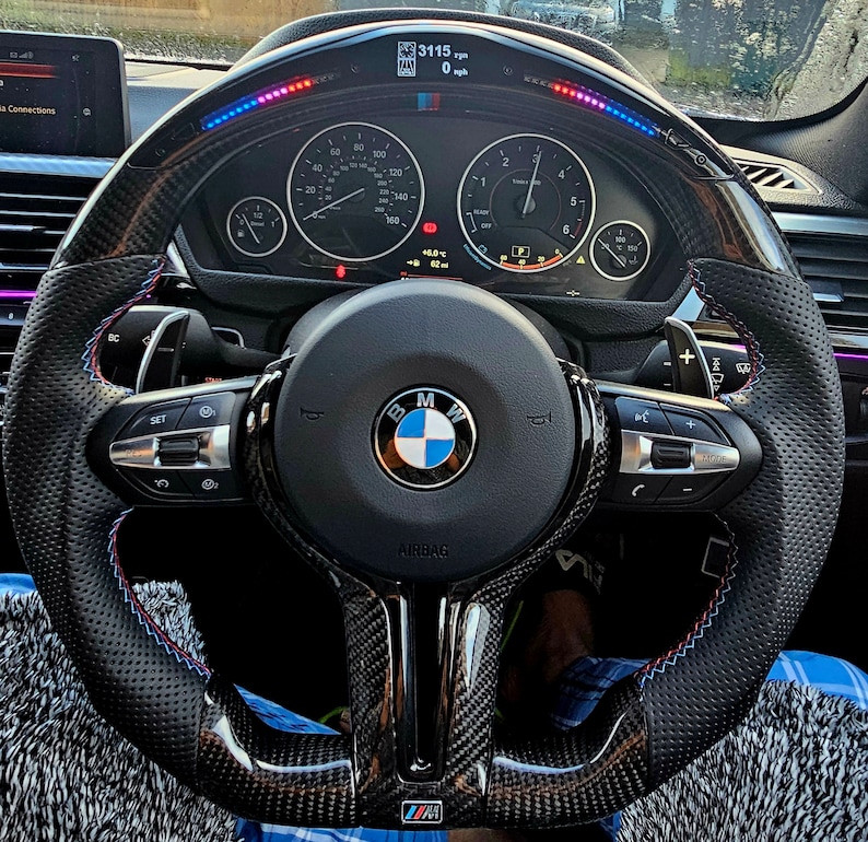 May include: A black leather steering wheel with a carbon fiber trim and blue and red stitching. The steering wheel has a BMW logo in the center and controls for the car's functions. The steering wheel is also equipped with a digital display that shows the car's speed, RPM, and other information.