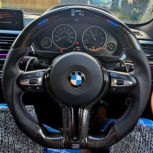 May include: A black leather steering wheel with a carbon fiber finish. The steering wheel has a BMW logo, red and blue stitching, and a digital display that reads "1833 rpm".