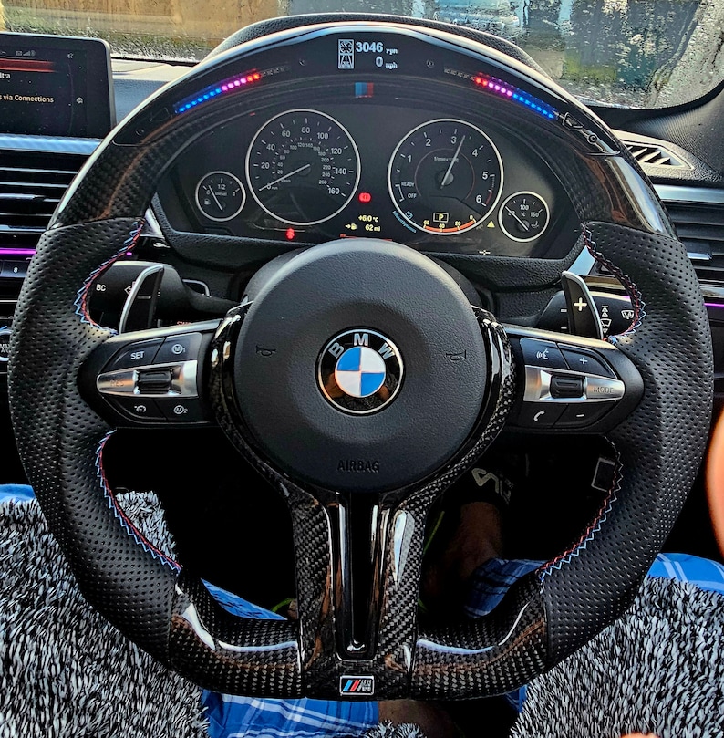 May include: A black leather steering wheel with a carbon fiber trim and blue and red stitching. The steering wheel has a BMW logo in the center and a digital display that reads "3046 rpm" and "0 mph".