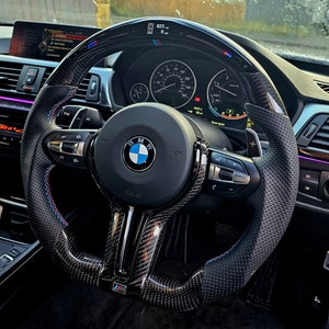 May include: A black leather steering wheel with a carbon fiber trim and blue and red stitching. The steering wheel has a BMW logo and controls for the car's functions. The steering wheel is in a car's interior.
