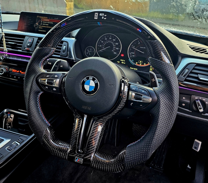 May include: A black leather steering wheel with a carbon fiber trim and blue and red stitching. The steering wheel has a BMW logo and controls for the car's functions. The steering wheel is in a car's interior.