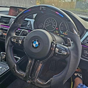 May include: A black carbon fiber steering wheel with blue, red, and white accents. The steering wheel has a perforated leather grip, paddle shifters, and a BMW logo in the center. The steering wheel also has a digital display that shows the car's speed and other information.