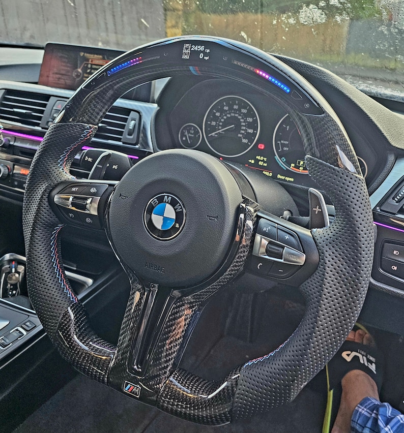 May include: A black carbon fiber steering wheel with blue, red, and white accents. The steering wheel has a perforated leather grip, paddle shifters, and a BMW logo in the center. The steering wheel also has a digital display that shows the car's speed and other information.