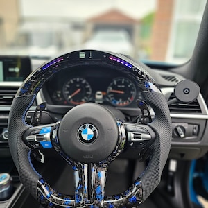K&ouml;nnte beinhalten: Ein schwarzes Lederlenkrad mit blauen und schwarzen Carbon-Akzenten. Das Lenkrad tr&auml;gt das BMW-Logo und den Schriftzug "M Performance" unten. Das Lenkrad verf&uuml;gt au&szlig;erdem &uuml;ber ein digitales Display mit einem blauen und violetten Lichtbalken.
