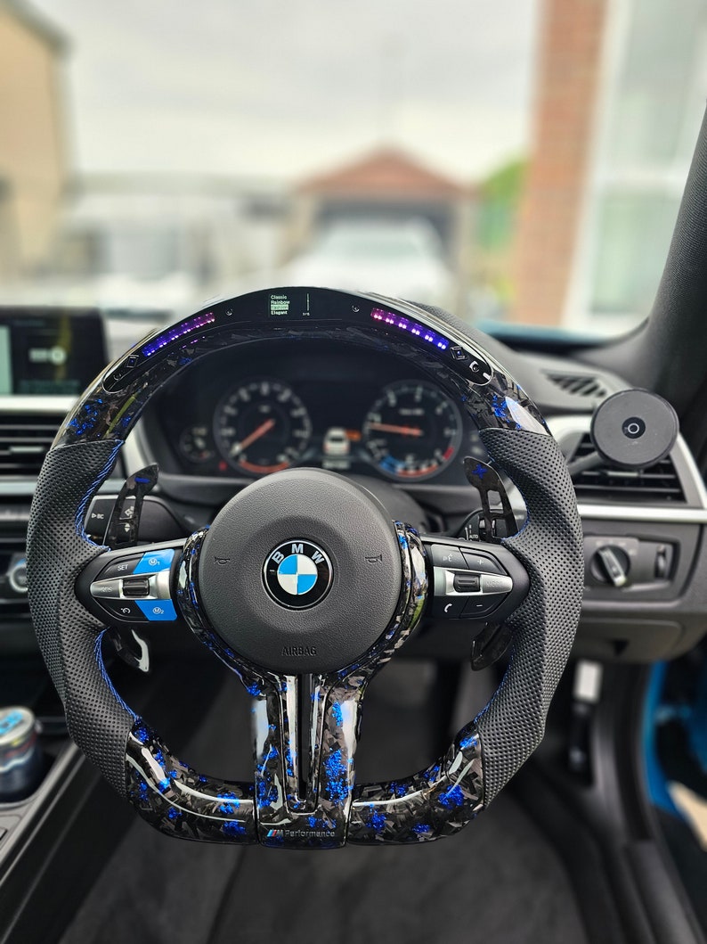 K&ouml;nnte beinhalten: Ein schwarzes Lederlenkrad mit blauen und schwarzen Carbon-Akzenten. Das Lenkrad tr&auml;gt das BMW-Logo und den Schriftzug "M Performance" unten. Das Lenkrad verf&uuml;gt au&szlig;erdem &uuml;ber ein digitales Display mit einem blauen und violetten Lichtbalken.