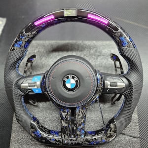 K&ouml;nnte beinhalten: Ein schwarzes Lederlenkrad mit blauen N&auml;hten und einem Karbonfaser-Muster. Das Lenkrad tr&auml;gt das BMW-Logo und den Schriftzug "Airbag". Es gibt mehrere Kn&ouml;pfe am Lenkrad, darunter eine Lautst&auml;rkeregelung, eine Telefontaste und eine Tempomat-Taste. Das Lenkrad hat auch eine Reihe von rosa LED-Leuchten oben herum.