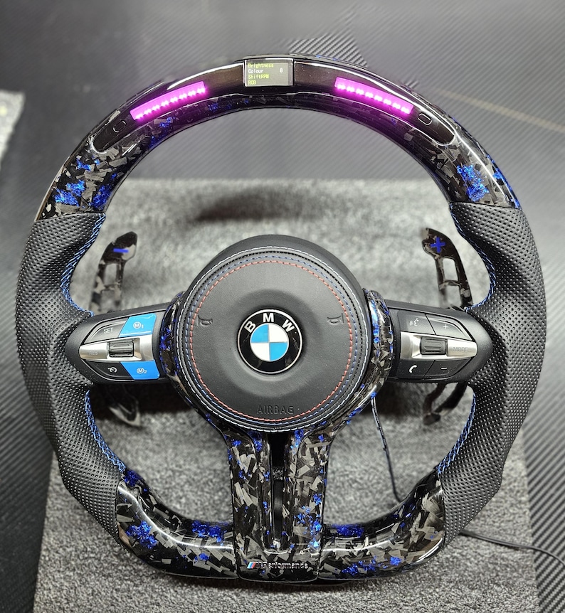 K&ouml;nnte beinhalten: Ein schwarzes Lederlenkrad mit blauen N&auml;hten und einem Karbonfaser-Muster. Das Lenkrad tr&auml;gt das BMW-Logo und den Schriftzug "Airbag". Es gibt mehrere Kn&ouml;pfe am Lenkrad, darunter eine Lautst&auml;rkeregelung, eine Telefontaste und eine Tempomat-Taste. Das Lenkrad hat auch eine Reihe von rosa LED-Leuchten oben herum.