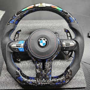 K&ouml;nnte beinhalten: Ein schwarzes Lederlenkrad mit blauen N&auml;hten und einem Karbonfaser-Muster. Das Lenkrad tr&auml;gt das BMW-Logo und den Schriftzug "Airbag". Das Lenkrad hat auch ein blaues und schwarzes Streifenmuster um den Rand.