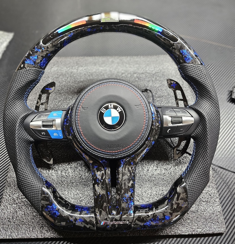 K&ouml;nnte beinhalten: Ein schwarzes Lederlenkrad mit blauen N&auml;hten und einem Karbonfaser-Muster. Das Lenkrad tr&auml;gt das BMW-Logo und den Schriftzug "Airbag". Das Lenkrad hat auch ein blaues und schwarzes Streifenmuster um den Rand.