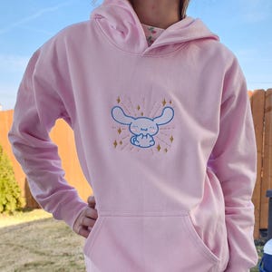 May include: A pink hoodie with a white embroidered character with blue eyes and a yellow starburst design.