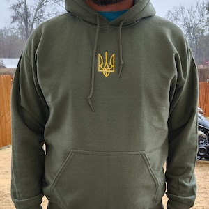 Puede incluir: Una sudadera con capucha verde con un símbolo de tridente ucraniano bordado en amarillo en la parte delantera.