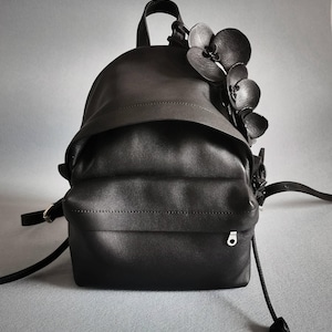 Pode incluir: Uma mochila de couro preto com um design floral na aba superior. A mochila tem um fecho de cordão e um pequeno laço de metal na frente.
