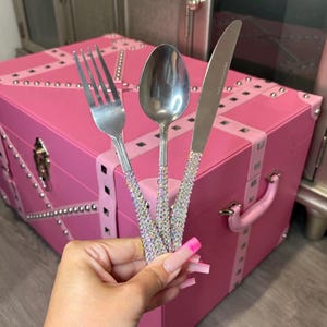 May include: A set of three stainless steel utensils consisting of a fork, a spoon, and a knife. Each handle is covered in small, iridescent, multi-coloured rhinestones that catch the light, providing a textured and reflective surface.