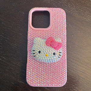 Puede incluir: Funda de tel&eacute;fono rosa cubierta de pedrer&iacute;a iridiscente. Presenta un dise&ntilde;o de Hello Kitty con un lazo rosa. El &aacute;rea de la lente de la c&aacute;mara est&aacute; delineada en rosa. La funda est&aacute; sobre una superficie de madera marr&oacute;n oscuro.