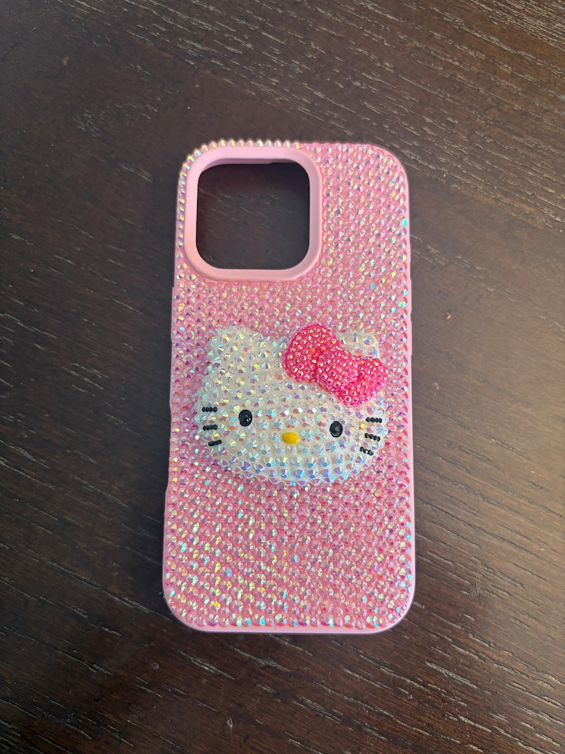 Puede incluir: Funda de tel&eacute;fono rosa cubierta de pedrer&iacute;a iridiscente. Presenta un dise&ntilde;o de Hello Kitty con un lazo rosa. El &aacute;rea de la lente de la c&aacute;mara est&aacute; delineada en rosa. La funda est&aacute; sobre una superficie de madera marr&oacute;n oscuro.