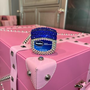 Könnte beinhalten: Ein blau verziertes Vaseline-Erdölgelee-Glas mit einem silbernen und blauen Kristall-Design. Das Glas steht auf einem rosa Koffer mit silbernen Nieten und Akzenten. Der Koffer hat einen rosa Lederriemen.