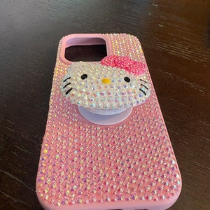 Puede incluir: Funda de tel&eacute;fono rosa cubierta de pedrer&iacute;a iridiscente, con un dise&ntilde;o de Hello Kitty adornado con pedrer&iacute;a, un lazo rosa y un agarre emergente blanco. La funda est&aacute; dise&ntilde;ada para proteger un tel&eacute;fono.