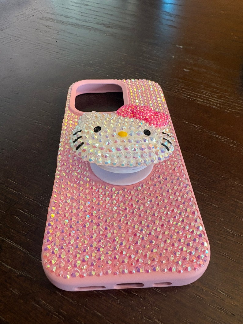 Puede incluir: Funda de tel&eacute;fono rosa cubierta de pedrer&iacute;a iridiscente, con un dise&ntilde;o de Hello Kitty adornado con pedrer&iacute;a, un lazo rosa y un agarre emergente blanco. La funda est&aacute; dise&ntilde;ada para proteger un tel&eacute;fono.