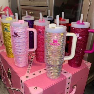 May include: A collection of bedazzled Stanley tumblers in various colors, including pink, gold, and purple. Each tumbler features a handle, a straw, and the Stanley logo. The tumblers are displayed on a pink trunk.