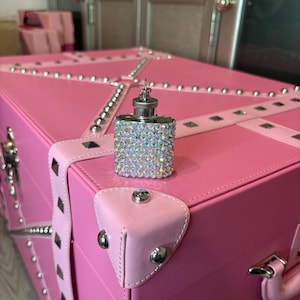Op de afbeelding: Een roze decoratieve kist met zilveren studs en een vierkante, met kristallen bedekte flacon. De kist heeft roze leren riemen en hoekbeschermers. De flacon heeft een zilveren dop en is bedekt met iriserende kristallen.