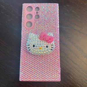 Puede incluir: Funda de tel&eacute;fono rosa cubierta de pedrer&iacute;a iridiscente. Presenta un dise&ntilde;o 3D de Hello Kitty con un lazo rosa. La funda tiene recortes para la c&aacute;mara y los botones del tel&eacute;fono.