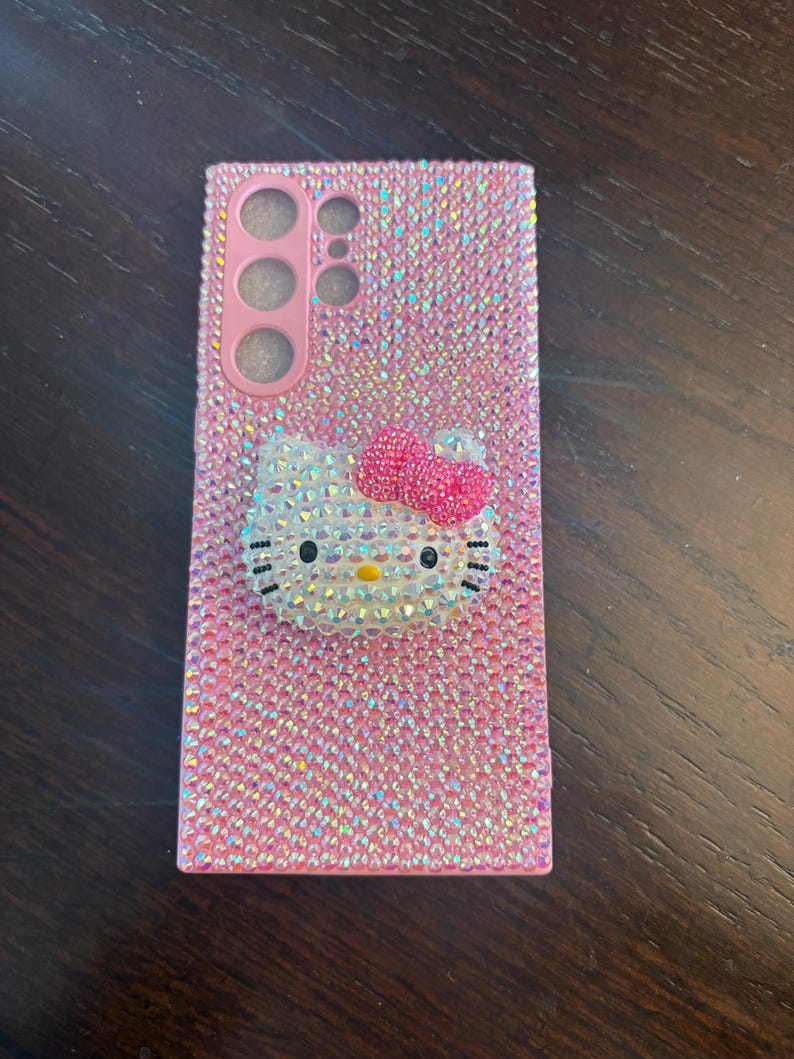 Puede incluir: Funda de tel&eacute;fono rosa cubierta de pedrer&iacute;a iridiscente. Presenta un dise&ntilde;o 3D de Hello Kitty con un lazo rosa. La funda tiene recortes para la c&aacute;mara y los botones del tel&eacute;fono.