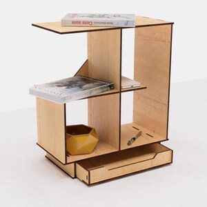May include: A wooden shelf unit with a drawer, featuring four compartments. The shelf unit is made of light wood with a natural finish. It is decorated with books, a small bowl, and a pen.