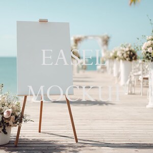 May include: A white easel with wooden legs stands on a wooden deck, with the text "EASEL MOCKUP" in white. The background features a beach wedding setup with floral arrangements and chairs, under a bright blue sky.